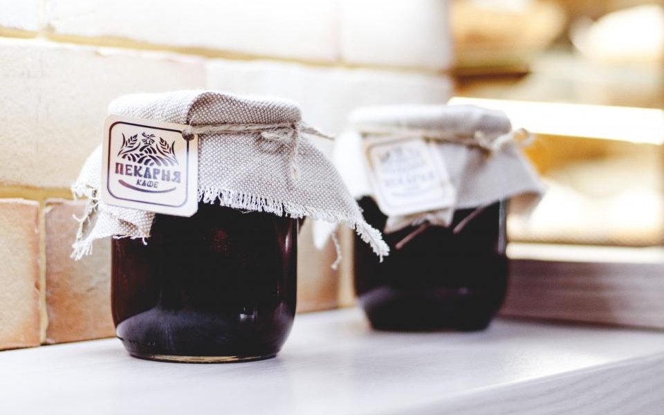 Two preserve jars with cloth-covered lids tied at the neck and small hand-written paper labels, standing on a shelf by a window