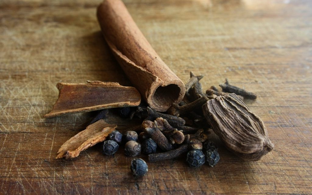 Whole spices on a dark wooden board — cinnamon quills, black cardamom, cloves and peppercorns