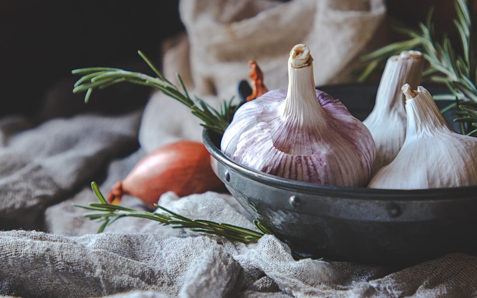 Whole garlic bulbs and a shallot in a battered metal bowl with rosemary sprigs on rumpled linen