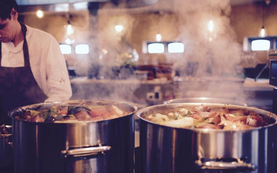A cook in a dark apron working behind two large stainless stockpots, steam rising in warm kitchen light