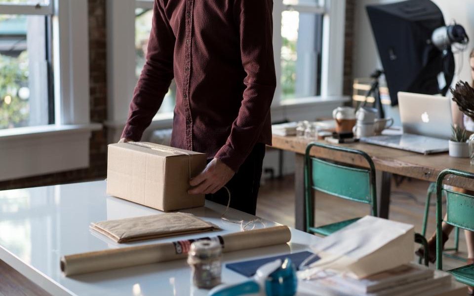 Someone closing a small cardboard parcel at a work table, beside a roll of kraft paper and tape