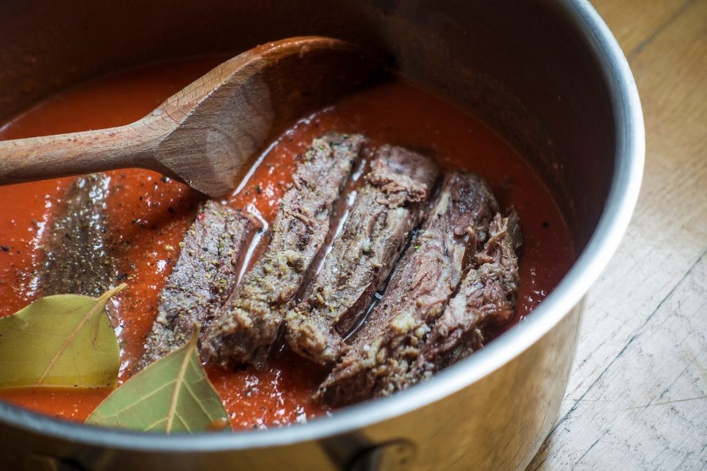Beef short ribs braising in deep red tomato sauce in a steel pot, a wooden spoon and bay leaves resting in it