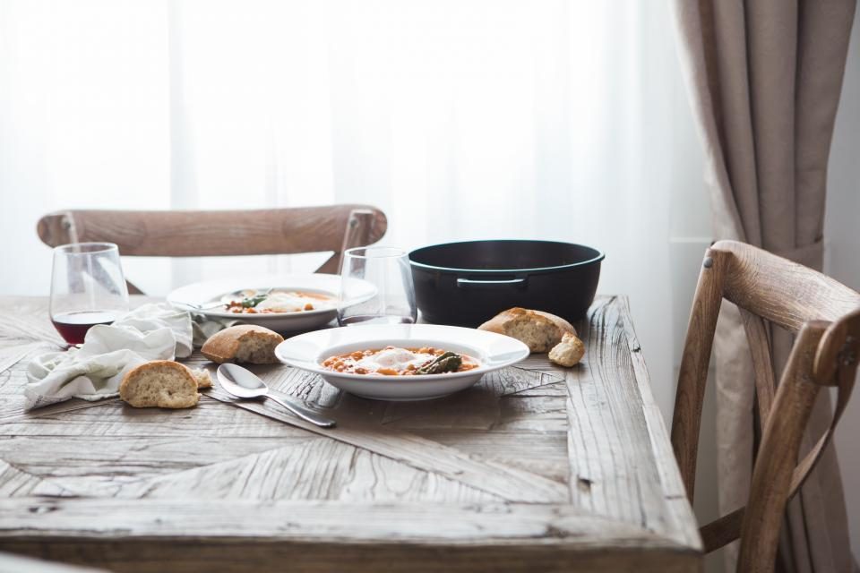 A rustic laid table with bread rolls, two bowls of soup, a cast-iron pot and a glass of red wine in soft daylight